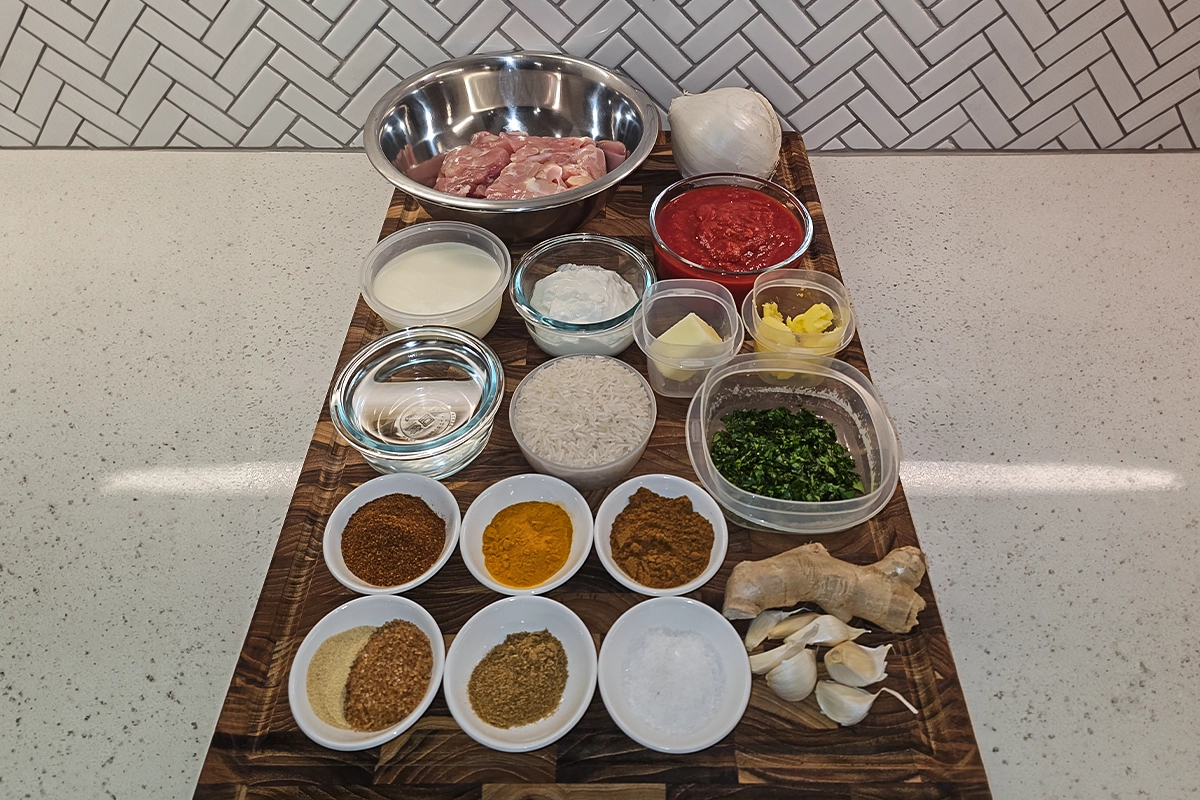 Top-down view of neatly arranged ingredients for Josh Cizek’s 30-minute butter chicken recipe, featuring raw chicken thighs, plain non-fat Greek yogurt, Kashmiri chili powder, turmeric, lemon juice, ground cumin, garam masala, kosher sea salt, finely grated raw ginger, diced white onion, minced garlic cloves, fenugreek powder, raw cane sugar, ghee, crushed tomatoes, heavy cream, unsalted butter, and fresh cilantro—set against a clean kitchen backdrop, ideal for a flavorful Indian curry preparation.