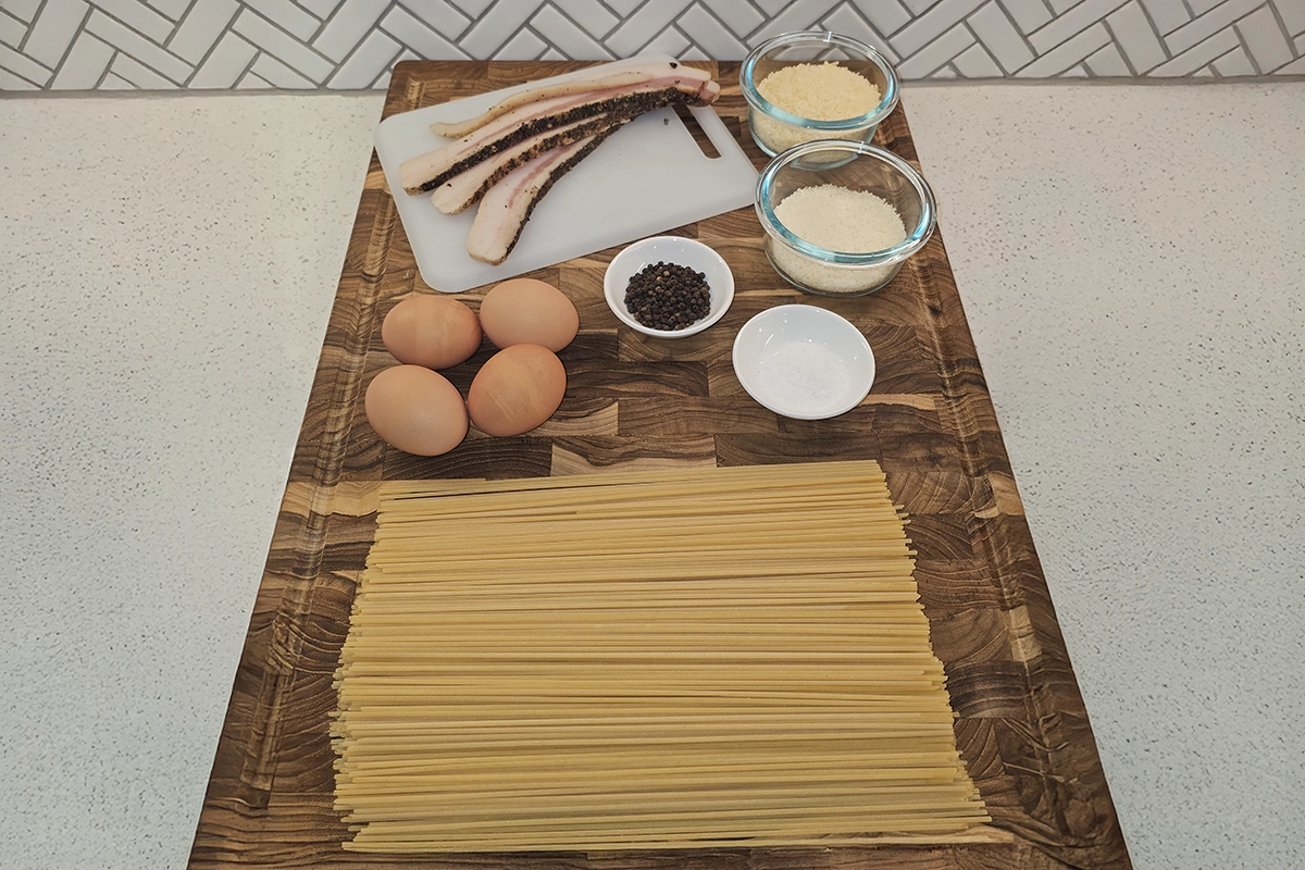 Top-down view of classic spaghetti carbonara ingredients arranged neatly on a wooden cutting board: uncooked spaghetti, four eggs, thick slices of guanciale, black peppercorns, salt, grated Pecorino Romano, and grated Parmigiano-Reggiano. Recipe by Josh Cizek Cooks.