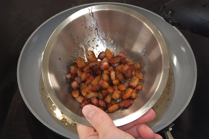 Close-up of a hand holding a stainless steel bowl filled with golden-brown, crispy rendered guanciale pieces, freshly cooked and glistening with fat over a stove. Recipe by Josh Cizek Cooks.